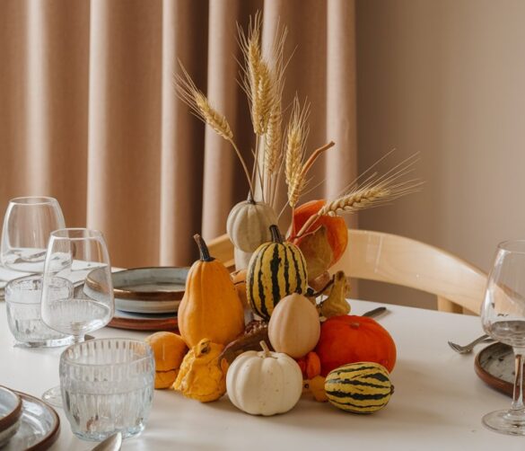 a-photo-of-a-fall-decorated-dining-table-there-s-a-JwZ8ryI4SPCZNUdHvHrdEg-_8iQsif_SJ-fRDaYVXXZbQ A photo of a fall-decorated dining table. There's a harvest-themed centerpiece made of mismatched gourds and wheat. The table is set for a meal, with plates, silverware, and glasses. The background has a beige curtain and a wooden shelf with a few items. The lighting is soft.