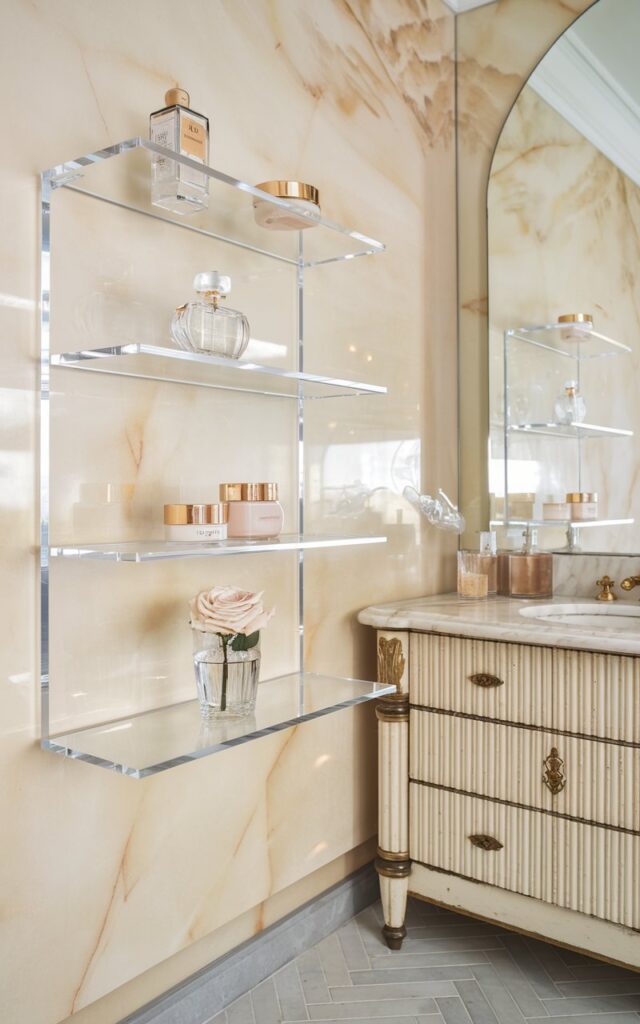 A photo of a European glam bathroom with crystal-clear Lucite shelves floating on a soft marble wall in pale champagne. The shelves hold delicate glass perfume bottles, gold-trimmed skincare jars, and a single rose in a slim crystal vase. Below, there's a vintage-inspired vanity with fluted details and brushed brass hardware. The flooring is herringbone tile. There's a large arched mirror in the room. The Lucite shelves blend into the background, letting the styling and soft-glow lighting speak volumes in understated sophistication.