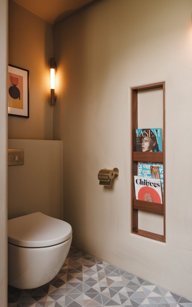 A photo of a mid-century modern bathroom with a built-in magazine rack. The bathroom has soft-toned walls, a curved-edge toilet, and geometric floor tiles. The magazine rack is seamlessly integrated into the wall beside the toilet. The rack has a slim, rectangular niche framed in walnut or teak wood, holding a few vintage-style magazines with bold, retro covers. The room has minimalist art, warm lighting, and brass or matte black fixtures.