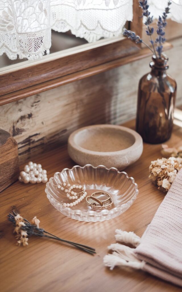A photo of a rustic cottagecore bathroom counter with a warm wood surface and soft natural textures. There's a small clear glass catch-all dish with a delicate, vintage-inspired edge holding a few dainty gold rings, pearl earrings, and floral hairpins. Nearby, there's a stoneware soap dish, a tiny dried flower bundle, and a fringed cotton hand towel. The backdrop features a wood-platted wall, an antique-style mirror, and a sprig of lavender in a repurposed glass bottle. Soft morning light filters through a lace curtain, creating a dreamy, nostalgic mood.