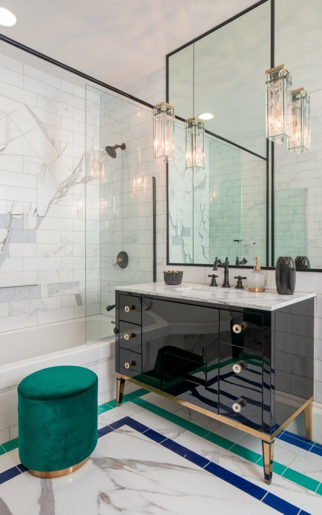 A photo of a chic Art Deco bathroom with white marble tiles featuring elegant gray veining. The tiles are accented by vibrant green and blue colored grout that highlights the geometric tile layout. The colored grout adds a playful yet sophisticated twist, making the marble's natural patterns pop with bold contrast. Glossy black fixtures with brass detailing, a sleek black vanity with mirrored accents, and a vintage-inspired emerald green velvet stool complete the luxe look. Bold geometric mirrors and crystal pendant lights enhance the glamorous vibe, blending classic Art Deco style with a fresh, modern chic edge.