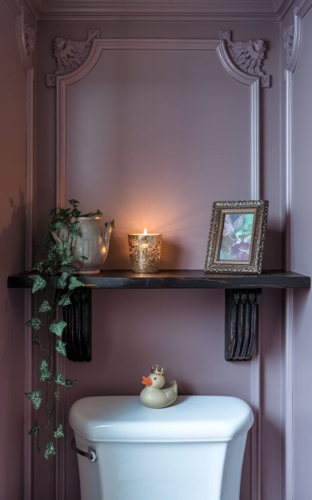 A photo of a cozy regal-style bathroom with a dark-stained wooden ledge mounted above a white toilet. The shelf holds a flickering gold-rimmed candle, a small trailing ivy plant in a ceramic pot, a tiny vintage art frame in muted jewel tones, and a whimsical rubber duck with a crown. The walls are painted in soft mauve, accented with ornate trim. The room is illuminated by natural light.