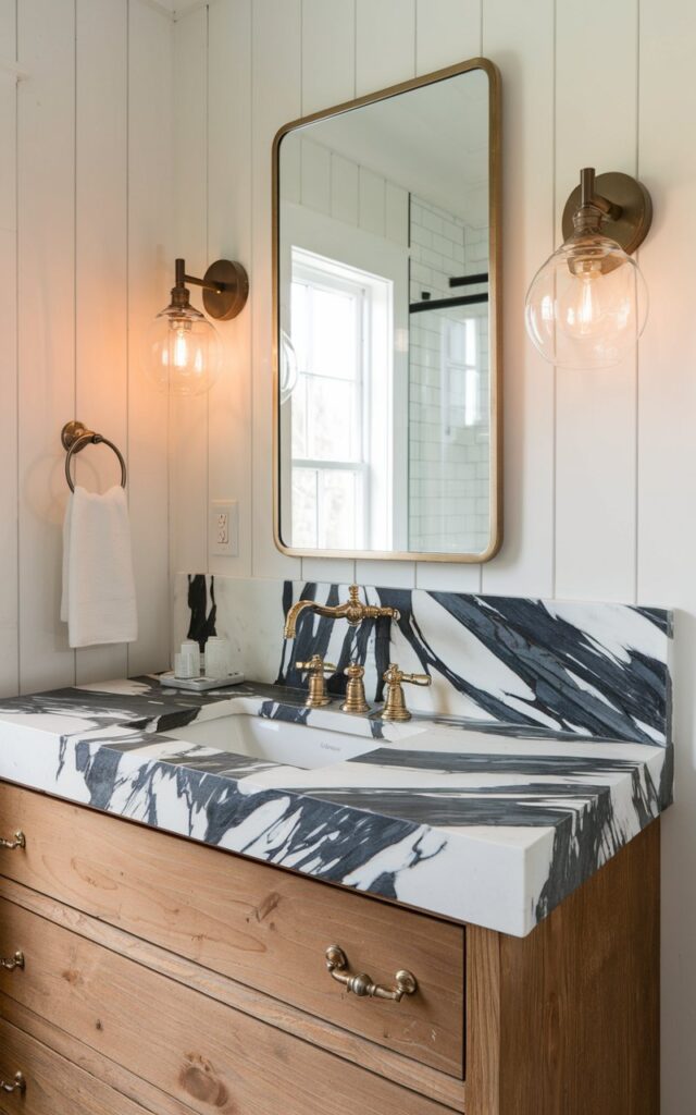A closer view of the marble slab countertop in the farmhouse-luxe bathroom. The marble is rich with bold gray and charcoal veining, creating a striking contrast against the raw wood tones. Vintage-style gold faucets and matching drawer pulls add a glamorous touch. The walls are covered in white vertical shiplap, and sconces with clear glass globes bring warm ambient light.