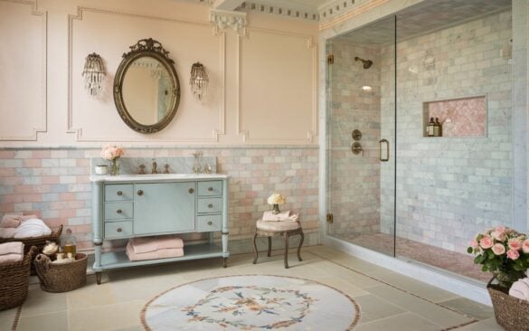 A photo of a fully furnished marble bathroom with a blend of English countryside charm and glamorous details. The vanity area features a soft pink-toned marble tile backsplash and a powder-blue vanity with antique brass fixtures. Above the vanity is an ornately framed oval mirror and crystal wall sconces. The flooring has a floral-patterned marble medallion at the center, bordered by pale stone tiles. The shower area is enclosed in clear glass and includes a recessed niche lined with pink marble. The walls are painted in a creamy blush hue with subtle paneling. The space has a vintage stool, fluffy towels, a vase of garden roses, and woven baskets. The atmosphere is classic and indulgently fresh.