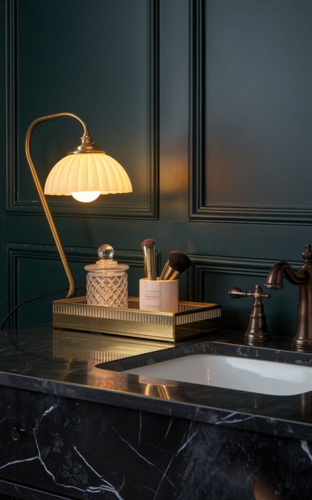 A photo of a Victorian + modern fusion bathroom vanity with a striking black marble countertop and brass fixtures, set against a panelled wall in deep green or navy. There is a clip-on small, stylish lamp with a curved brass neck and a milk-glass or pleated shade, on the edge of the vanity, glowing softly with warm yellow light. The lamp casts a cozy ambiance over the counter, which holds a mix of vintage and modern elements—cut-glass jars, a velvet-trimmed tray, and a sleek gold makeup brush holder. The blend of ornate details and clean lines creates a space that’s moody, refined, and perfectly layered.