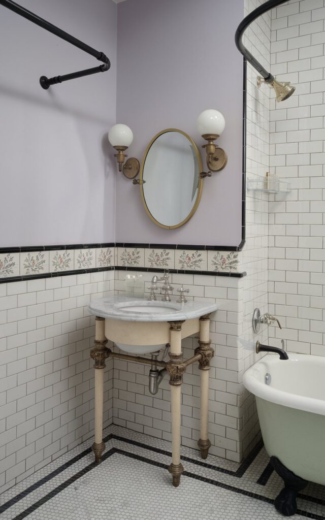 A photo of a small modern Victorian bathroom with a corner sink mounted on a compact marble-topped vanity with ornate legs and brushed gold hardware. The walls are lined with white subway tiles and feature a decorative floral border tile. Above the sink, a petite oval mirror with a vintage brass frame is flanked by twin globe sconces. The space also features a clawfoot tub with a curved shower curtain rail, black-and-white mosaic floor tiles, and soft wall colors like pale lavender. The overall vibe is refined, space-efficient, and classically charming.