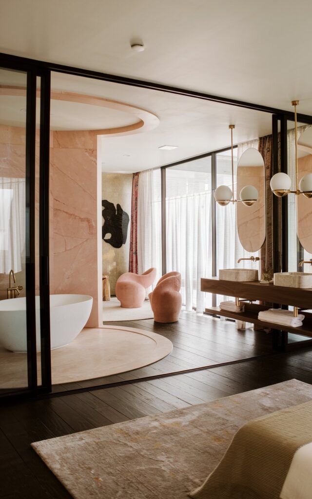 A photo of a chic mid-century modern bedroom with an open-plan ensuite. The bedroom features a dark-stained wood flooring and a plush area rug. There's a freestanding oval tub near the sleeping area, framed by a curved partial wall clad in blush pink marble. Gold-accented globe pendants hang over a walnut floating vanity with double sinks and oval mirrors. The shared space includes sculptural lounge seating, abstract wall art, and a touch of velvet in accessories. Large sliding glass doors bring in soft natural light, while sheer curtains add privacy without breaking flow.