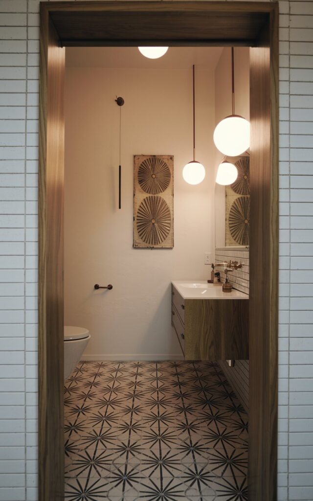 A photo of a mid-century modern bathroom with a teak-finished doorway. The bathroom has a floating wood vanity, globe pendant lighting, and a patterned tile floor with sunburst motifs. There is a vintage wall art piece and minimalist brass accents. The lighting is soft, ambient, and even.
