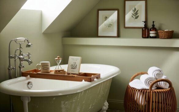 A photograph of a chic, spa-like bathroom blending modern and cottagecore aesthetics. A freestanding, antique clawfoot tub with a matte white finish is the central focus, positioned beneath a soft skylight that casts diffused natural light. A rustic wooden bath tray rests across the tub’s rim, displaying a "Lavender Chamomile" ceramic mug, a small sand timer, and a vintage copy of "Pride and Prejudice," while a plush, pebbled bath mat sits invitingly nearby. The sage green walls are adorned with two framed botanical prints, and a rattan basket overflowing with rolled white towels and amber glass dispensers sits neatly against a floating shelf, completing the serene and inviting atmosphere.