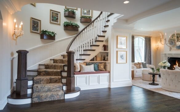 A photo of a fully furnished posh English countryside glam style living room. The room features a grand, statement-making staircase with natural stone treads and rich wood railings. The staircase incorporates Moroccan patterned risers, side-mounted sconces, and LED strip lights. A reading nook and built-in storage drawers are located beneath the staircase. The side wall has a gallery wall of framed art. Plant shelves line the railing, and the base of the staircase is anchored by a dramatic oversized newel post. The living room is lavishly decorated with plush seating, a roaring fireplace, layered rugs, and gold-accented décor, exuding both countryside charm and glamorous sophistication.
