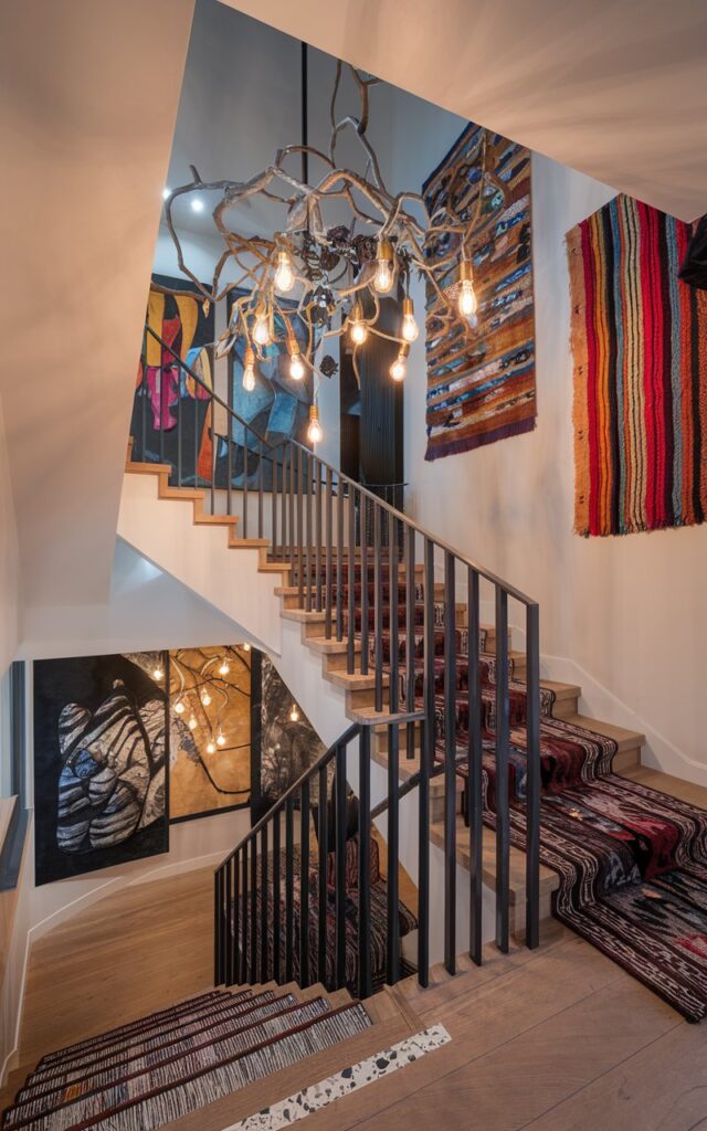 A photo of a modern eclectic home interior with a dramatic sculptural chandelier mounted above the staircase landing. The chandelier features an artistic arrangement of intertwined metal branches with exposed Edison bulbs. The staircase has natural wood and terrazzo treads and sleek black railings. Bold artwork, patterned rugs, and colorful textiles surround the landing, adding layers of personality. Soft, ambient lighting accentuates the eclectic mix of styles, textures, and colors, making the space feel vibrant and curated.