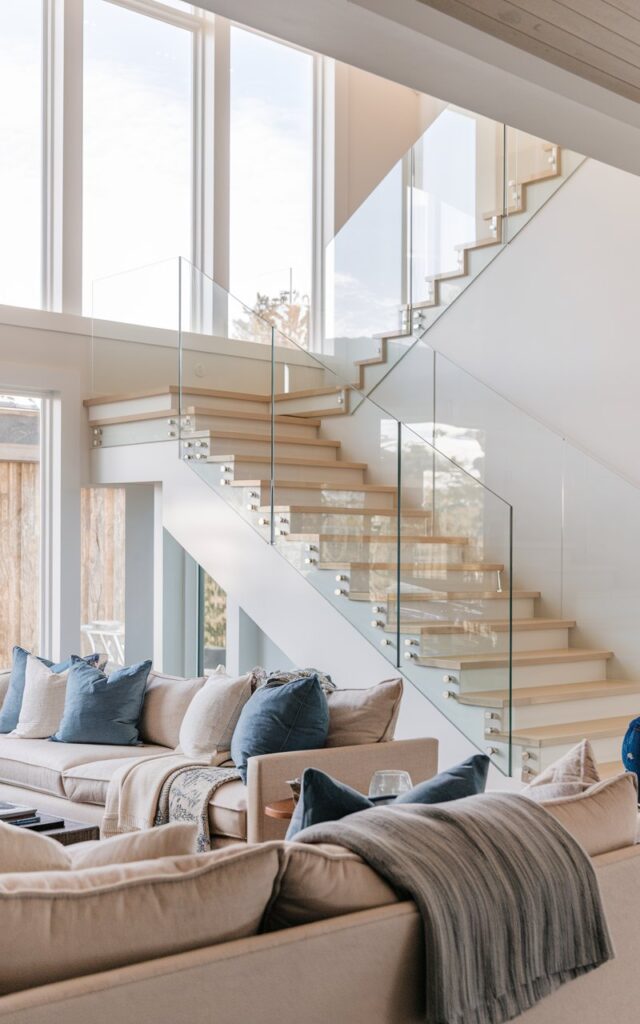 A photo of a living room with an alpine chic and coastal style. There's a floating staircase with sleek glass railings. The stairs have light-toned wooden treads and are supported by a hidden stringer. The glass panels are frameless and reflect soft natural light from large windows. The living room is fully furnished with plush sofas, layered throws, woven textures, and a neutral palette accented with ocean blues. Natural light is soft.