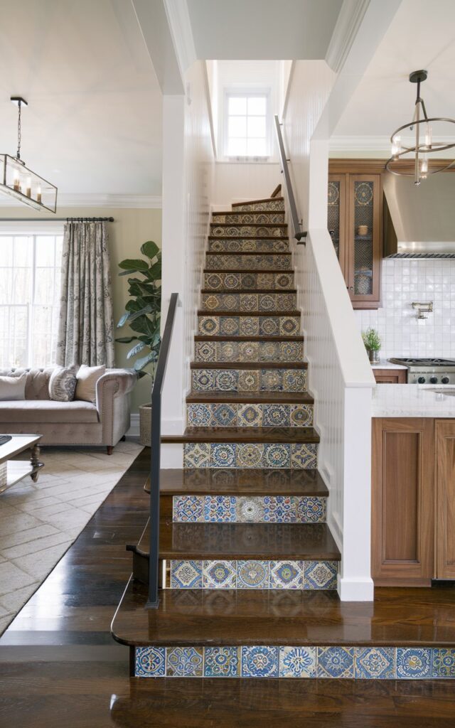 A photo of a modern-traditional home interior with a fully furnished living room and kitchen. The living room has a plush sofa, a coffee table, and a potted plant. The kitchen has wooden cabinetry, a stove, and a sink. The sleek stairs are positioned between the living room and kitchen, with vibrant Moroccan patterned tiles on the risers. The stair treads are polished dark wood, and there's a simple black metal railing. The lighting fixtures in both spaces are elegant.