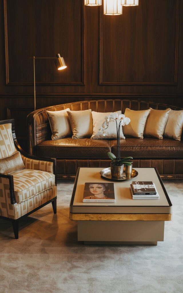 A photograph of a luxurious Art Deco living room, centered around a plush, dark brown leather couch with intricately stitched detailing and generously scattered cream-colored throw pillows. The couch is complemented by a patterned armchair angled towards it, featuring bold cream and gold geometric shapes and a single decorative lumbar pillow. A sleek rectangular coffee table with gold accents and a glass top displays "The Kiss" by Klimt, a silver tray with a single white orchid, and a stack of art books, bathed in the soft glow of a brass floor lamp and a geometric chandelier. The room's walls are adorned with dark wood paneling and a subtle, textured wallpaper, creating a sense of classic elegance.