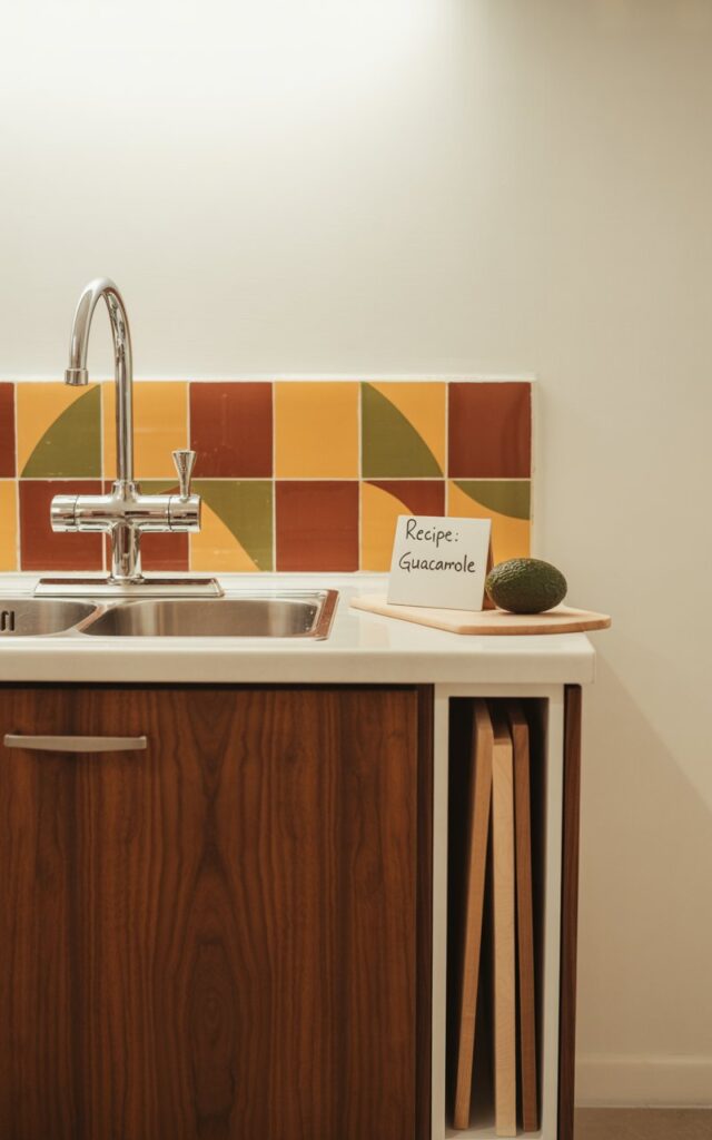 A photograph of a pristine mid-century modern kitchen counter, the focal point being a sleek walnut cabinet containing a brushed stainless steel sink. A vibrant geometric backsplash in warm terracotta, ochre, and olive green tones provides a striking contrast behind the sink, while a vertical wooden cutting board is neatly housed within a narrow cabinet slot to the right. Resting on the counter beside the cutting board is a single, ripe avocado next to a small, handwritten sign that reads "Recipe: Guacamole". Soft, diffused natural light gently illuminates the clean lines and earthy textures of the kitchen, casting long, subtle shadows across the walnut grain and highlighting the warm colors of the backsplash.