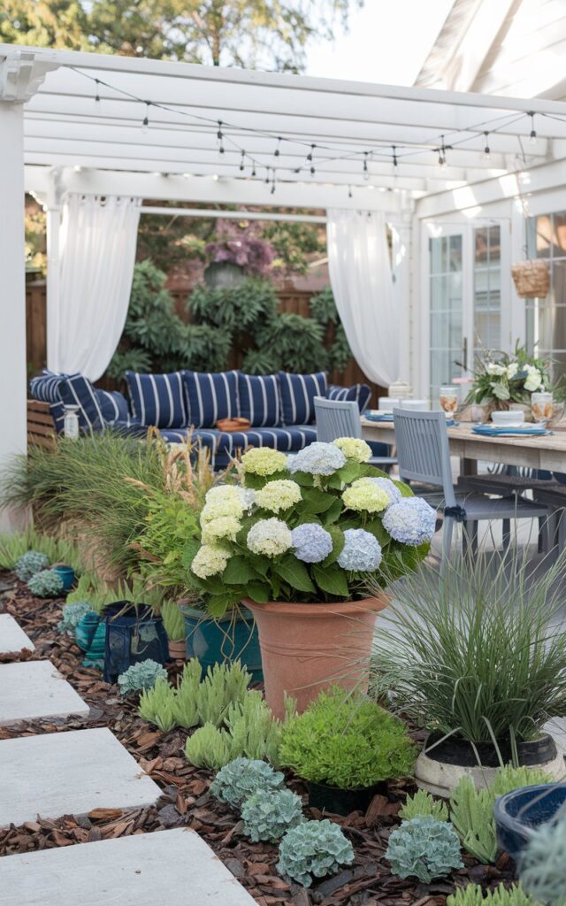 A wide view of a charming Americana and coastal-style backyard. There are potted plants of various sizes and heights, filled with flowering hydrangeas, grasses, ferns, and dwarf palms, artfully grouped along the backyard's edges. The ground is layered with creeping thyme, lamb's ear, and mulch, adding visual depth and coastal texture. In the background, a white pergola shades a navy-striped lounge set, and a rustic wooden dining table is set with coastal-themed dinnerware. Breezy white curtains sway in the breeze as string lights drape overhead.