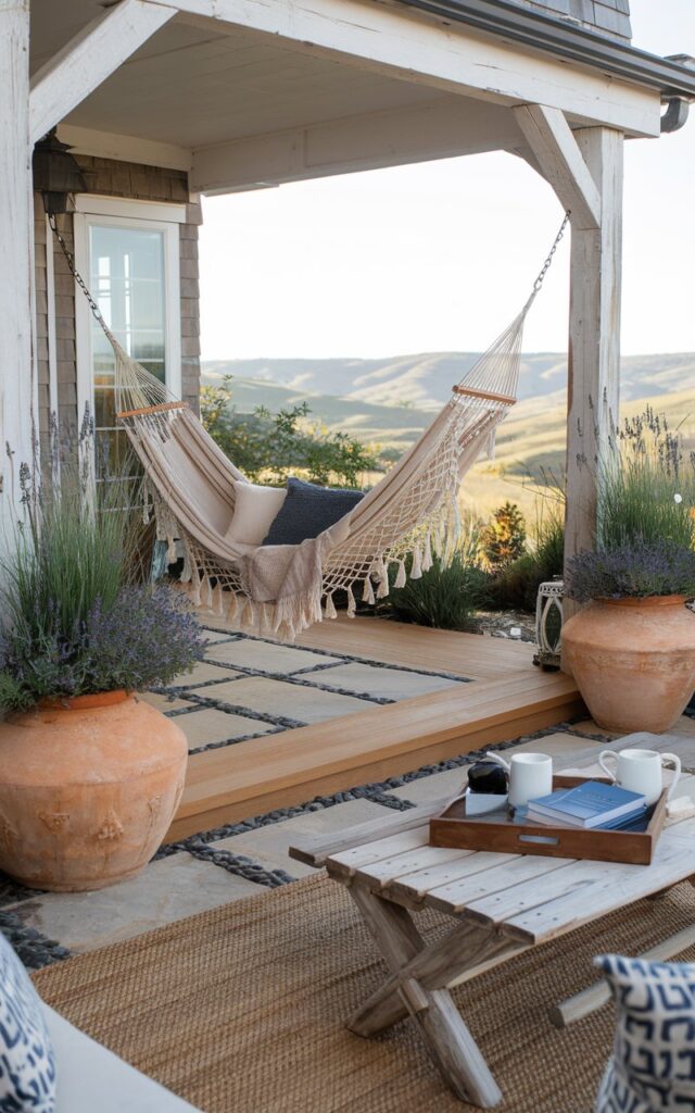 A photo of a coastal-alpine chic backyard patio attached to a house. The patio features a hammock strung between two sturdy, whitewashed wooden posts. The patio floor is a mix of natural stone and warm-toned decking. A cozy throw blanket and oversized knit cushions rest in the hammock, inviting relaxation. Surrounding the space are large terracotta pots filled with lavender, grasses, and small evergreens. A driftwood coffee table sits nearby with a tray of books and hot cocoa mugs. The backdrop showcases panoramic views of rolling hills, giving the space a serene, getaway vibe.