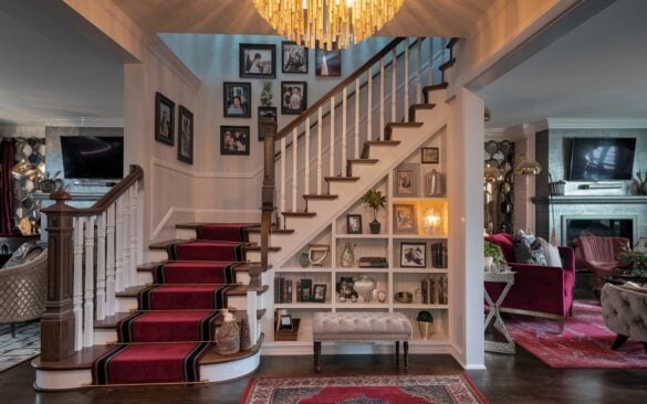 A photo of a fully furnished living room with a staircase at night. The staircase is the centerpiece, with polished wooden treads and a bold, contrasting runner. The balusters are patterned, and there's an intricately carved newel post. Beneath the stairs, there's a cozy mini-library with neatly arranged books, small plants, and decorative objects. A small upholstered bench sits near the landing. The staircase wall has a gallery of family photos in varied vintage-style frames. Above, a dramatic statement chandelier casts a warm, golden glow over the stairs. The living room has velvet cushions, a fireplace with a TV, metallic accents, and layered rugs, blending rustic charm with glamorous sophistication. The overall scene feels warm, inviting, and visually dynamic, capturing the perfect balance of functionality and decorative flair.