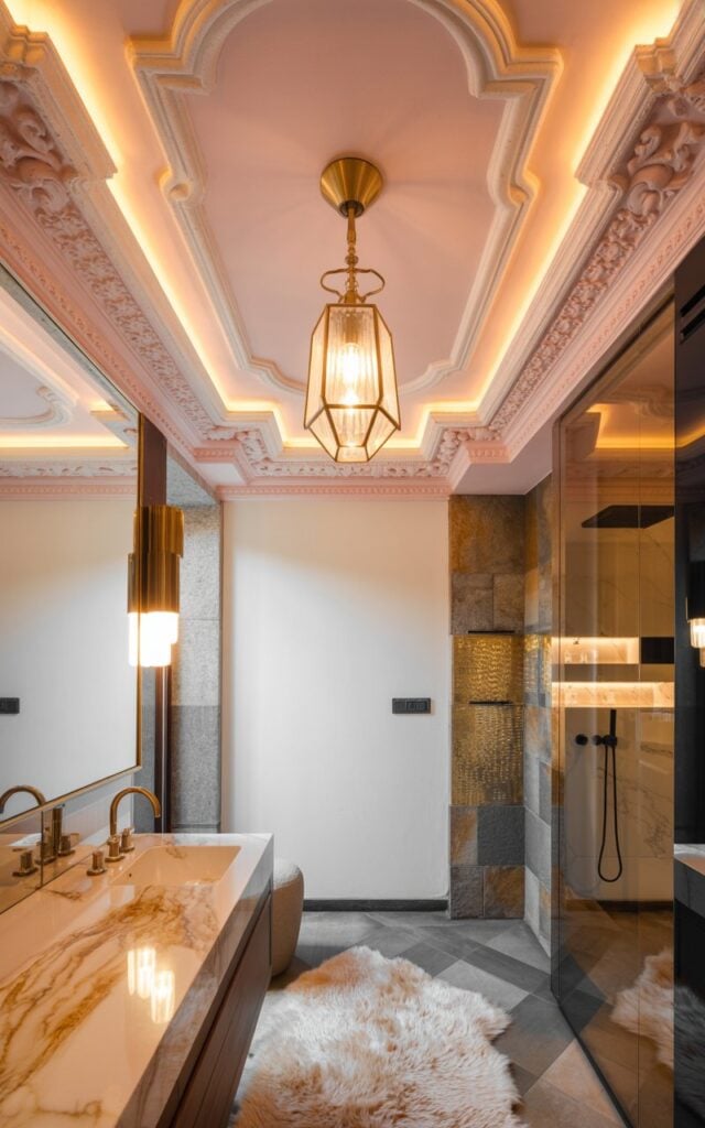 A luxurious bathroom interior photograph showcasing a glamorous alpine chic design, with an ornate soft blush pink ceiling as the stunning focal point, featuring intricate crown molding and decorative plasterwork in warm undertones. An elegant brass pendant light with geometric crystal accents hangs prominently from the center, casting a golden ambient glow that illuminates the ceiling's romantic hue and architectural details, while marble countertops with rich gold veining and polished brass fixtures create a cohesive metallic palette. The space is grounded by natural stone accent walls in warm gray tones, a plush cream faux fur rug, and a glass-enclosed shower area with sleek frameless doors. Warm LED strip lighting hidden within the molding and strategically placed sconces create layers of sophisticated illumination, perfectly balancing opulent glamour with the cozy, refined atmosphere of a high-end mountain retreat.