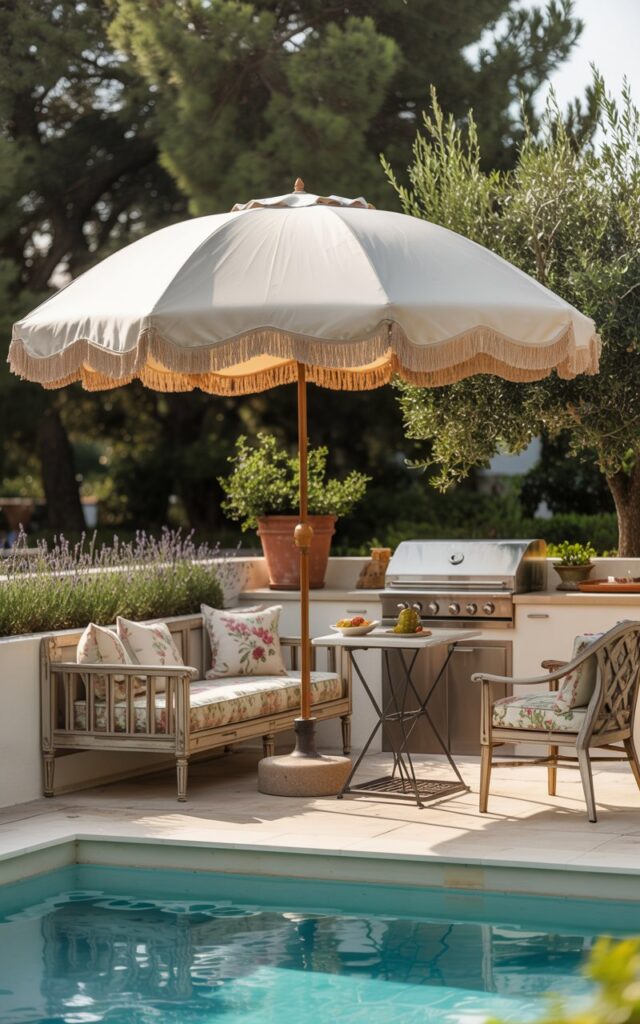 A photograph of a serene shabby chic patio overlooking a sparkling turquoise pool. A large, cream-colored oversized umbrella with delicate fringe details gracefully floats above a comfortable lounge seating area featuring distressed wooden chairs with floral patterned cushions. A small bistro table sits adjacent to a compact outdoor cooking station with stainless steel appliances, while potted lavender and olive trees add pops of color. Soft, golden hour light bathes the patio, creating a relaxed and inviting atmosphere.