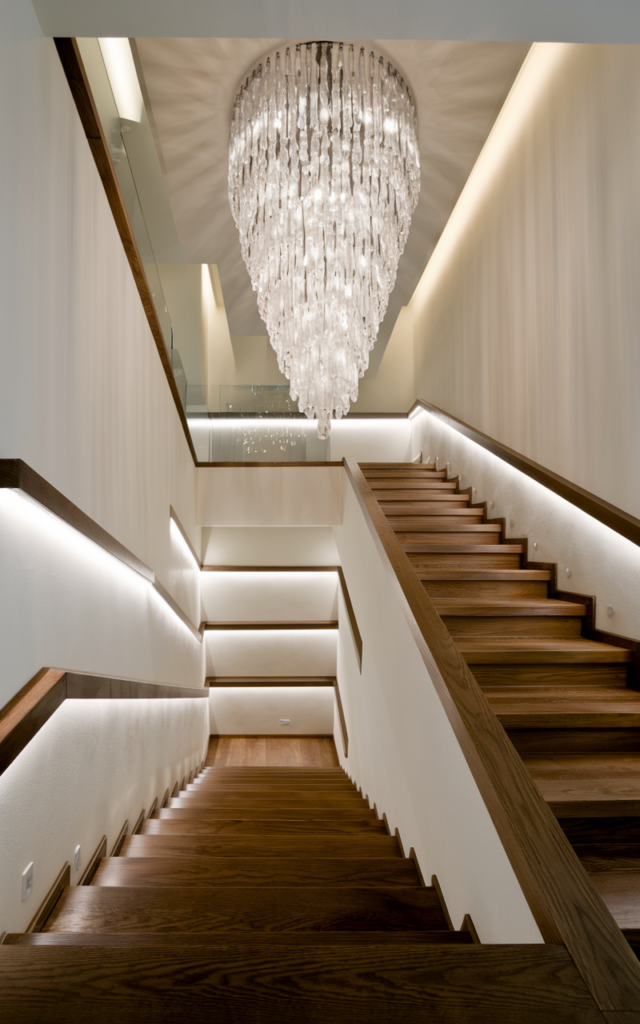 An elegant and natural-looking realistic photograph of a staircase featuring a dramatic cascading chandelier above paired with subtle recessed step lights along the staircase wall, creating layered lighting from top to bottom, decorated in modern luxury home decor style with natural lighting