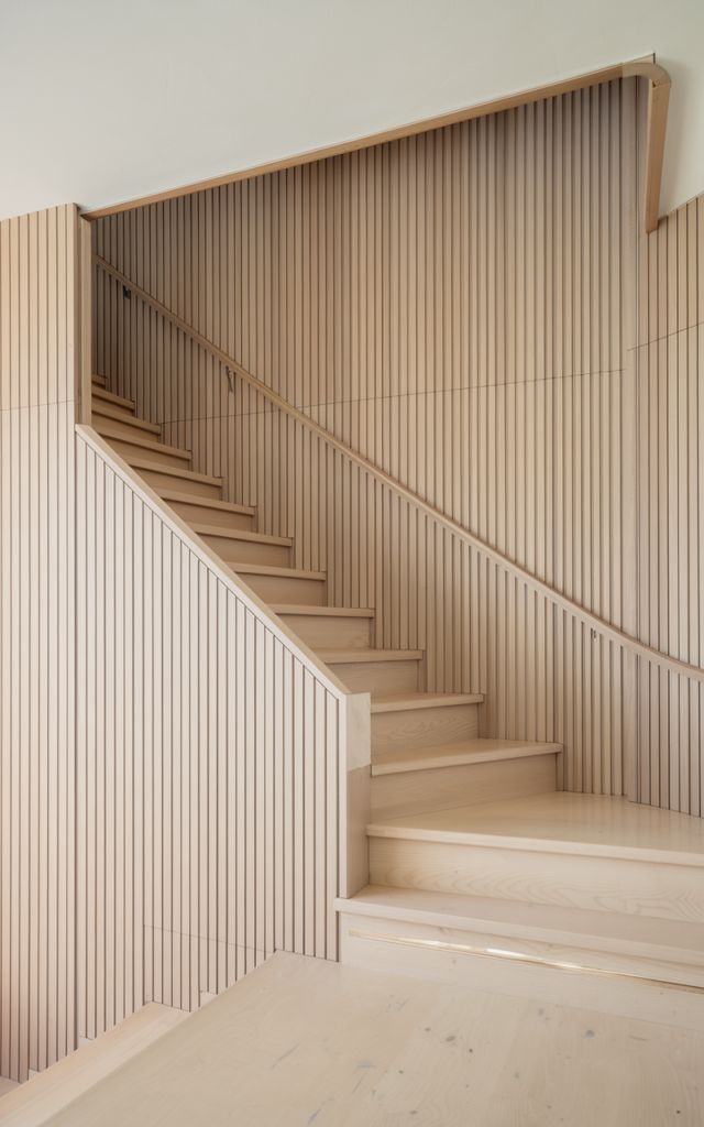 An elegant and natural-looking realistic photograph of a staircase wall covered in slim vertical slat molding panels from floor to ceiling beside a light wood staircase decorated in Scandinavian modern home decor style with natural lighting