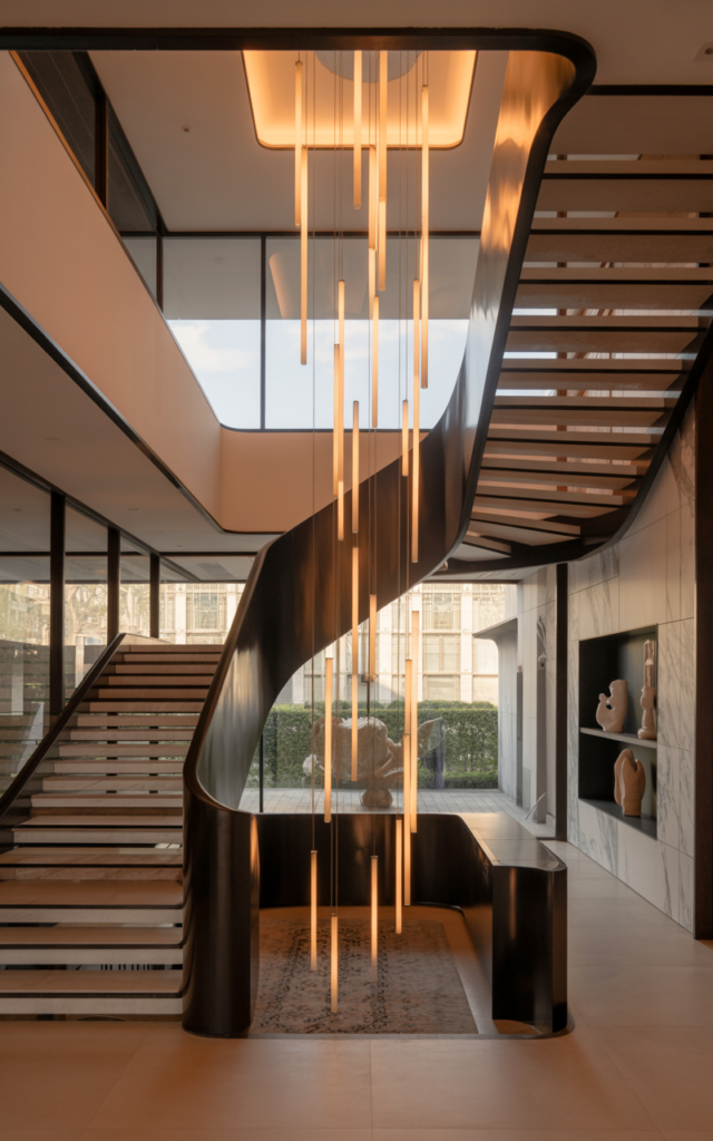 An elegant and natural-looking realistic photograph of a tall entry staircase with long vertical pendant lights hanging dramatically through a double-height stairwell beside the staircase, decorated in modern luxury home decor style with natural lighting