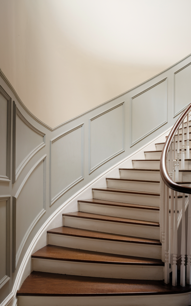 An elegant and natural-looking realistic photograph of a curved staircase foyer enhanced with subtle molding details tracing the stair’s movement along the wall painted in a soft neutral palette decorated in refined classic style with natural lighting