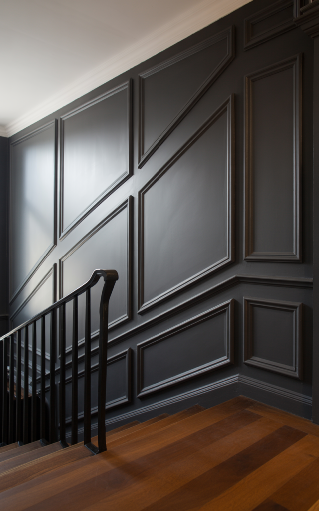 An elegant and natural-looking realistic photograph of a staircase accent wall featuring oversized molding panels painted in deep charcoal beside a staircase with black railing decorated in modern luxury home decor style with natural lighting
