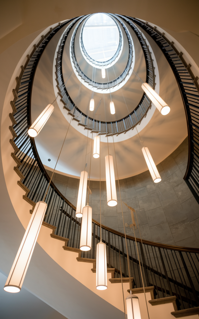 An elegant and natural-looking realistic photograph of a circular staircase highlighted by multiple pendant lights hanging at different heights following the spiral path in a tall stairwell decorated in modern contemporary interior style with natural lighting