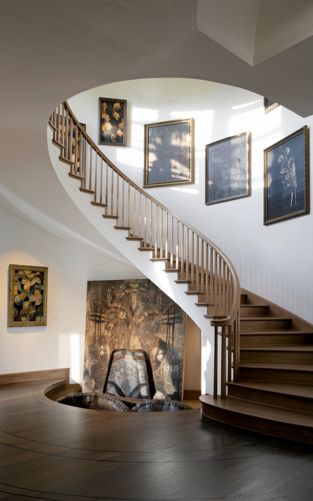 An elegant and natural-looking realistic photograph of a curved staircase foyer anchored by an oversized statement artwork at the base of the stairs balancing the sweeping architectural lines decorated in gallery-inspired contemporary style with natural lighting