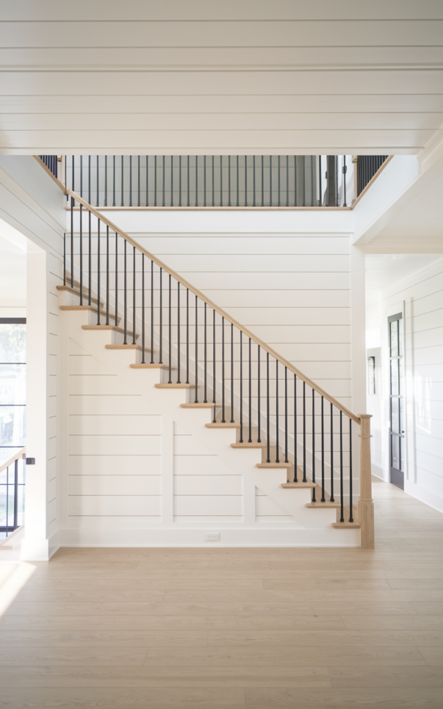 An elegant and natural-looking photograph of a minimalist staircase featuring slim black balusters, white walls, and light wood treads in a bright open foyer decorated in modern farmhouse style with natural lighting