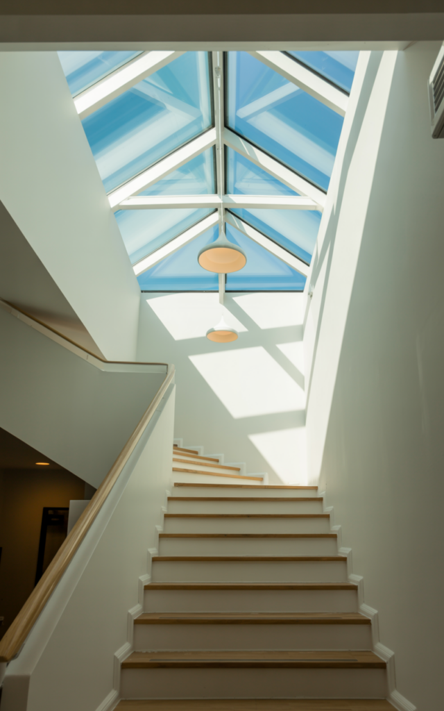 An elegant and natural-looking realistic photograph of a staircase positioned beneath a large skylight that floods the stairwell with daylight while a suspended pendant light hangs centrally for nighttime illumination, decorated in modern airy interior style with natural lighting