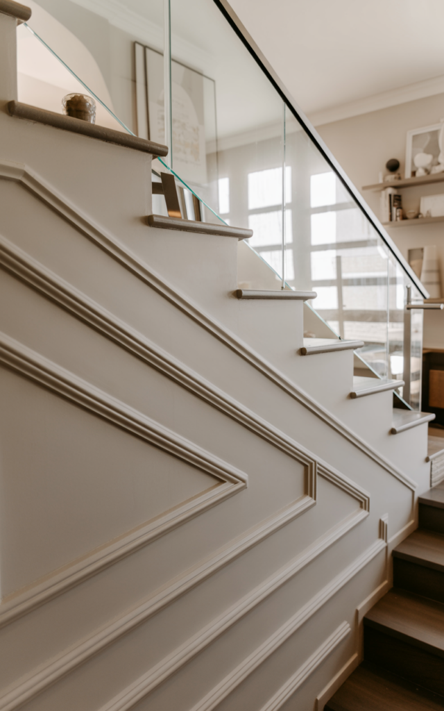 An elegant and natural-looking realistic photograph of a staircase wall with horizontal molding panel lines stretching across the stairwell beside a modern staircase with glass railing decorated in contemporary home decor style with natural lighting