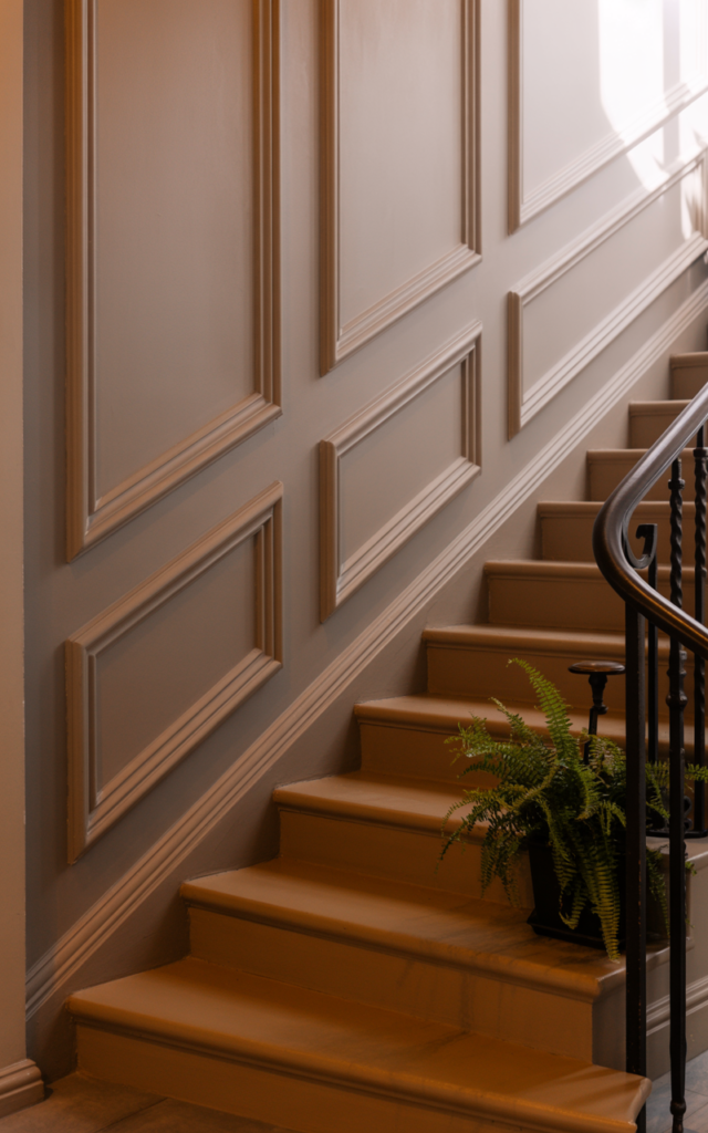 An elegant and natural-looking realistic photograph of a staircase wall adorned with subtle picture frame molding painted the same soft neutral shade as the wall above warm wood steps decorated in elegant transitional style with natural lighting