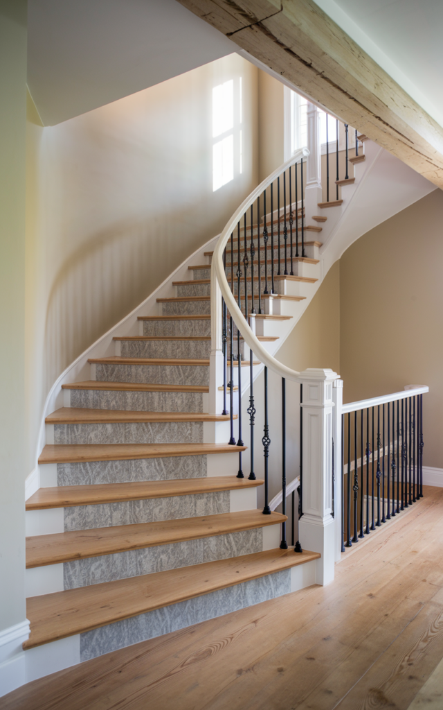 An elegant and natural-looking photograph of a staircase with white risers painted in a subtle muted gray stencil pattern, natural wood treads, and black iron railing decorated in modern farmhouse style with natural lighting