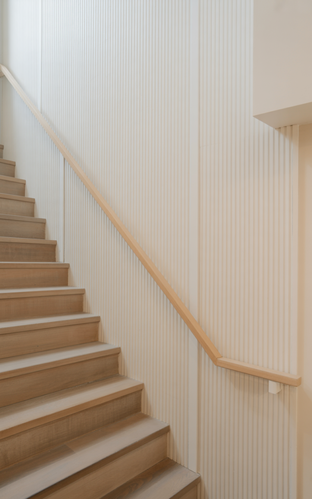 An elegant and natural-looking realistic photograph of a staircase wall covered in vertical fluted molding panels adding subtle ridged texture beside a light wood staircase with minimalist railing decorated in Scandinavian home decor style with natural lighting
