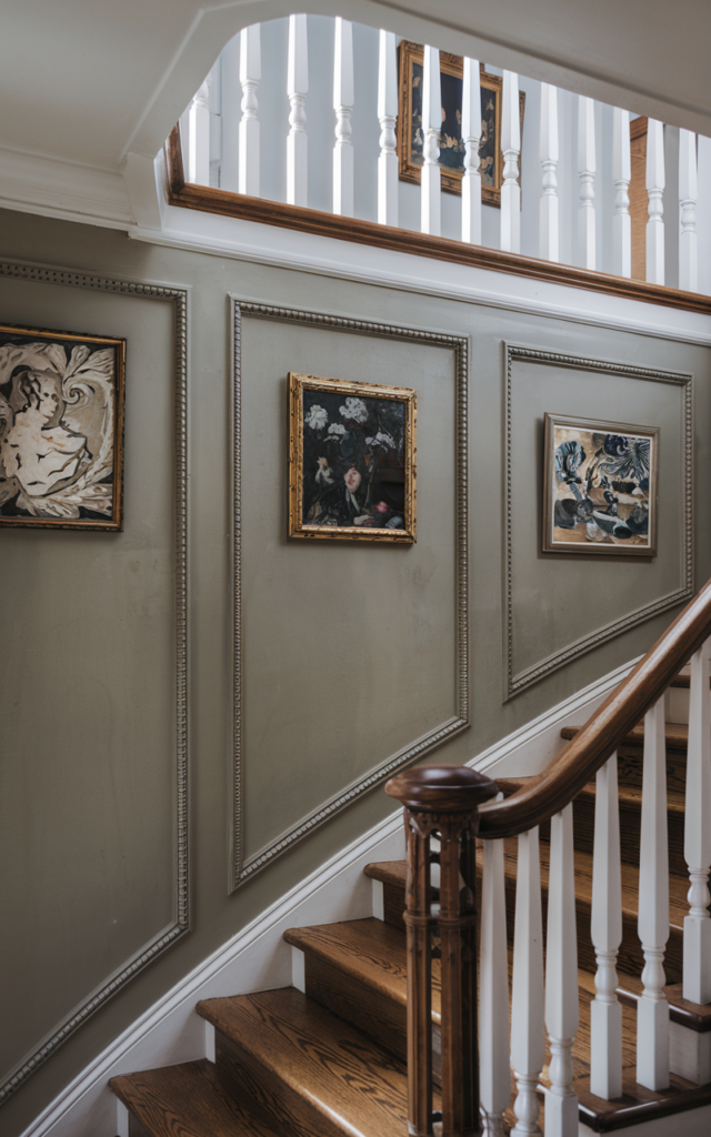 An elegant and natural-looking realistic photograph of a staircase landing wall framed with decorative molding panels containing elegant artwork above a wooden staircase with white balusters decorated in transitional home decor style with natural lighting