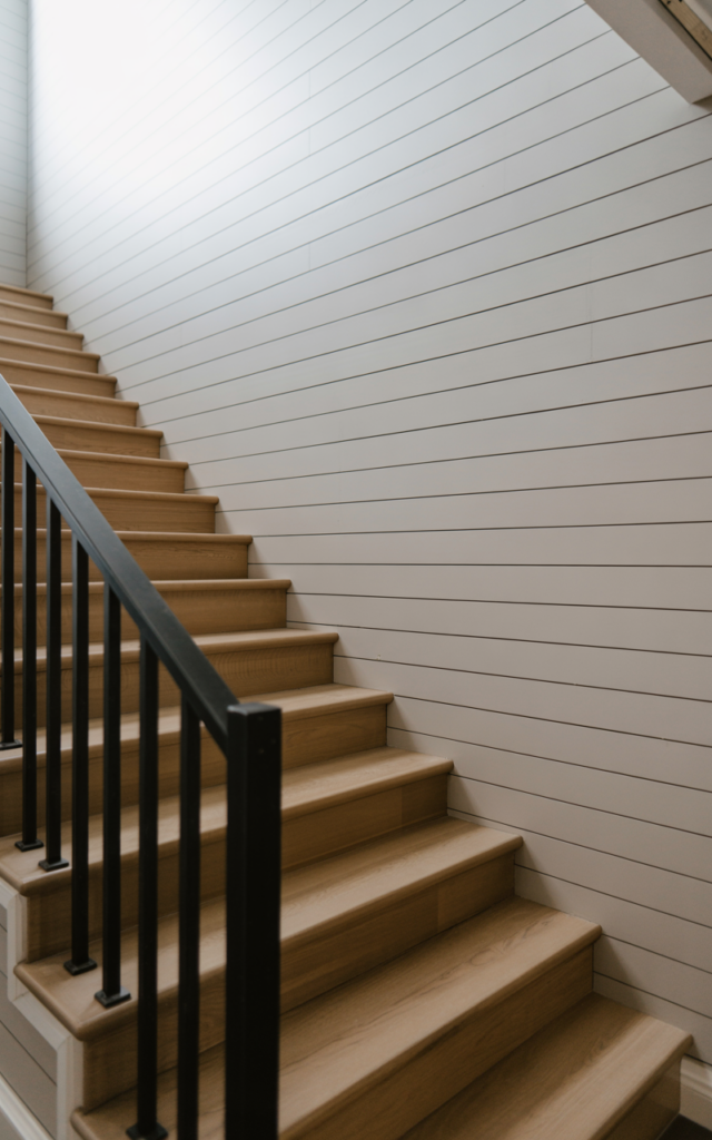 An elegant and natural-looking photograph of a staircase with vertical white shiplap accent wall climbing upward alongside light oak steps and black metal railing decorated in modern farmhouse style with natural lighting