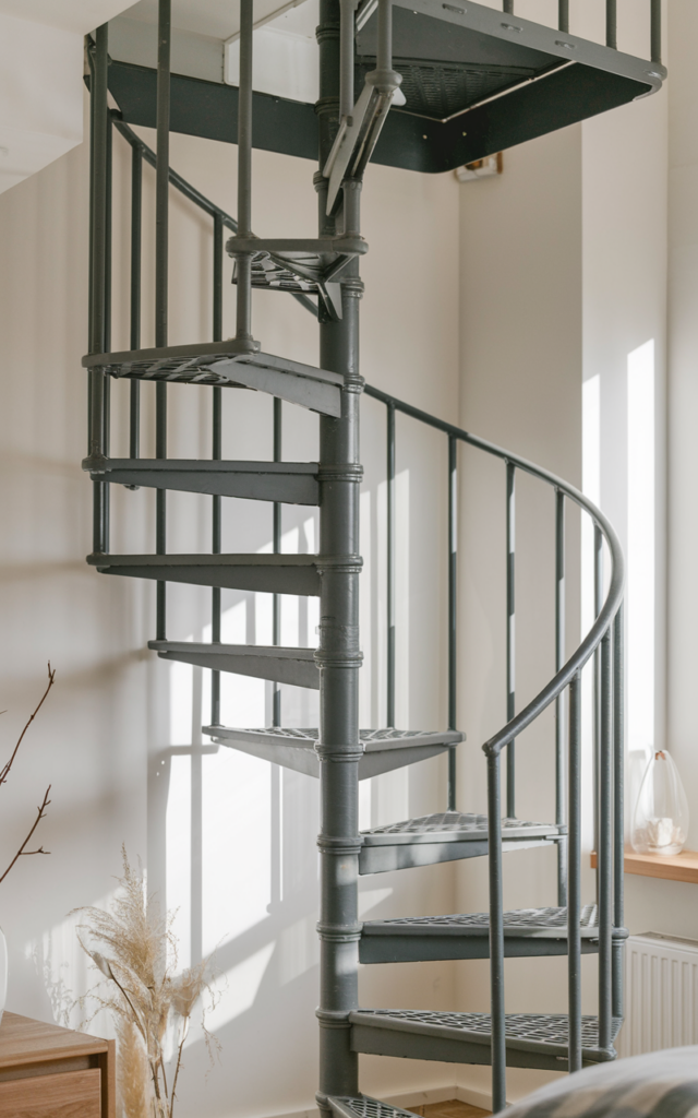 An elegant and natural-looking realistic photograph of a compact spiral staircase neatly installed in the corner of a small loft apartment with slim railings and open risers maximizing space efficiency decorated in minimalist urban interior style with natural lighting