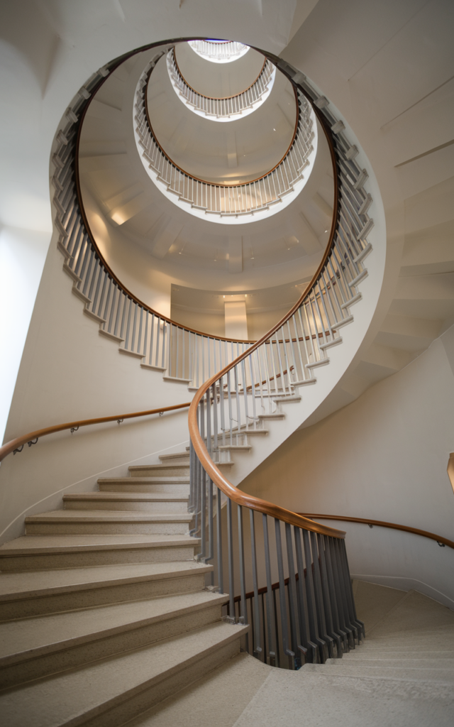 An elegant and natural-looking realistic photograph of a spiral staircase with a smoothly curved wooden handrail following the circular form of the stairs with metal balusters and a tall open stairwell decorated in modern architectural home decor style with natural lighting