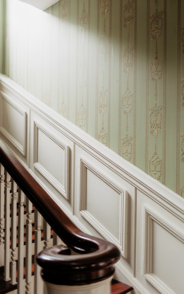 An elegant and natural-looking realistic photograph of a staircase wall enhanced with classic white wainscoting panels, dark wood banister and soft heritage wall color above the molding decorated in timeless Victorian style with natural lighting