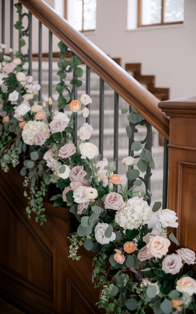 An elegant and natural-looking realistic photograph of a staircase railing adorned with elegant flower clusters of roses, hydrangeas, and ranunculus attached every few steps with delicate greenery accents decorated in soft romantic floral wedding style with natural lighting