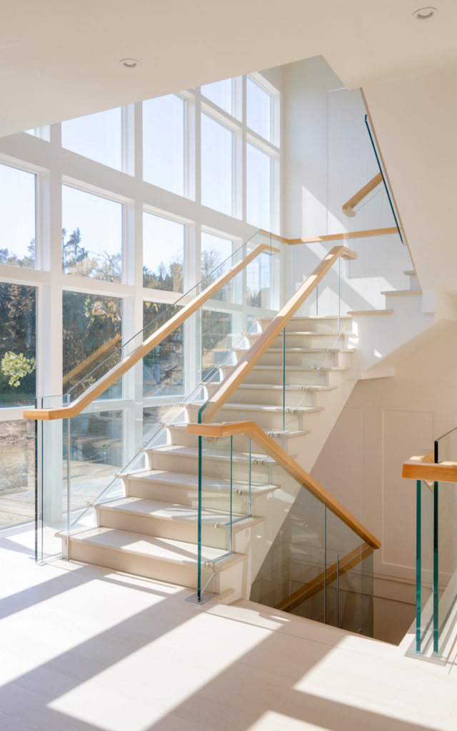 An elegant and natural-looking realistic photograph of a staircase with clear glass railing panels topped with a warm wooden handrail beside a bright open stairwell with sunlight pouring through large windows decorated in contemporary home decor style with natural lighting