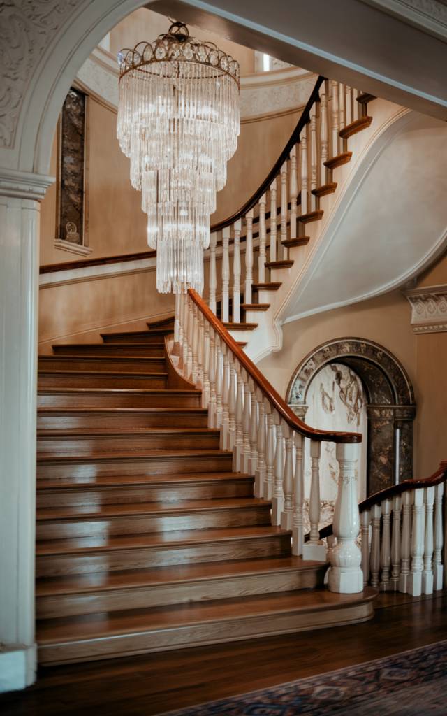 An elegant and natural-looking realistic photograph of a tall staircase with a cascading crystal chandelier hanging above polished wood steps and detailed railing creating a dramatic focal point decorated in grand Victorian style with natural lighting