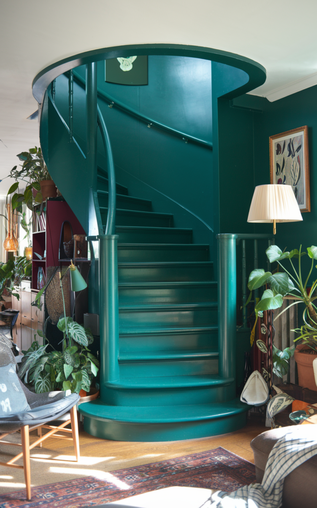An elegant and natural-looking realistic photograph of a bold circular staircase painted in deep emerald green with smooth spiral railings acting as a statement feature in a stylish living room decorated in eclectic modern interior style with natural lighting