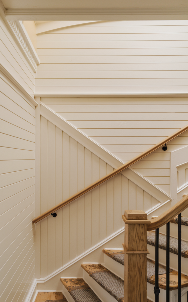 An elegant and natural-looking realistic photograph of a staircase wall finished with board and batten paneling painted in a soft warm white, contrasting lightly painted upper wall and simple wood handrail decorated in modern farmhouse style with natural lighting