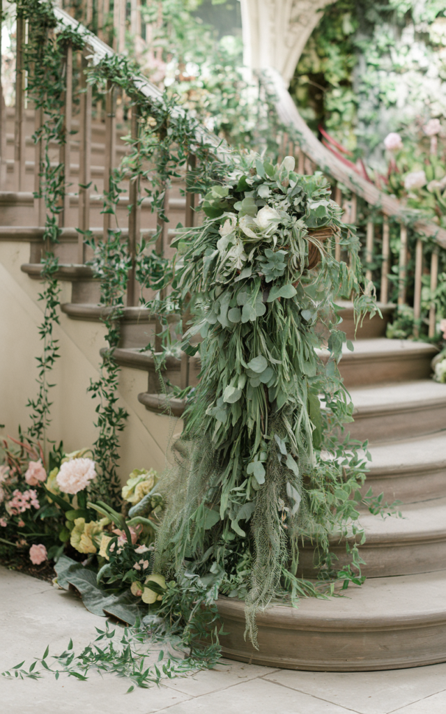 An elegant and natural-looking realistic photograph of a staircase decorated with cascading ivy, eucalyptus, and ruscus greenery spilling down from the banister like a botanical waterfall along the railings decorated in whimsical garden wedding style with natural lighting