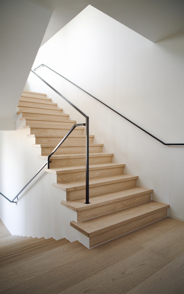 An elegant and natural-looking realistic photograph of a modern staircase with a slim matte black square-profile metal handrail mounted along a white wall with light oak steps and minimal decor decorated in modern minimalist home decor style with natural lighting