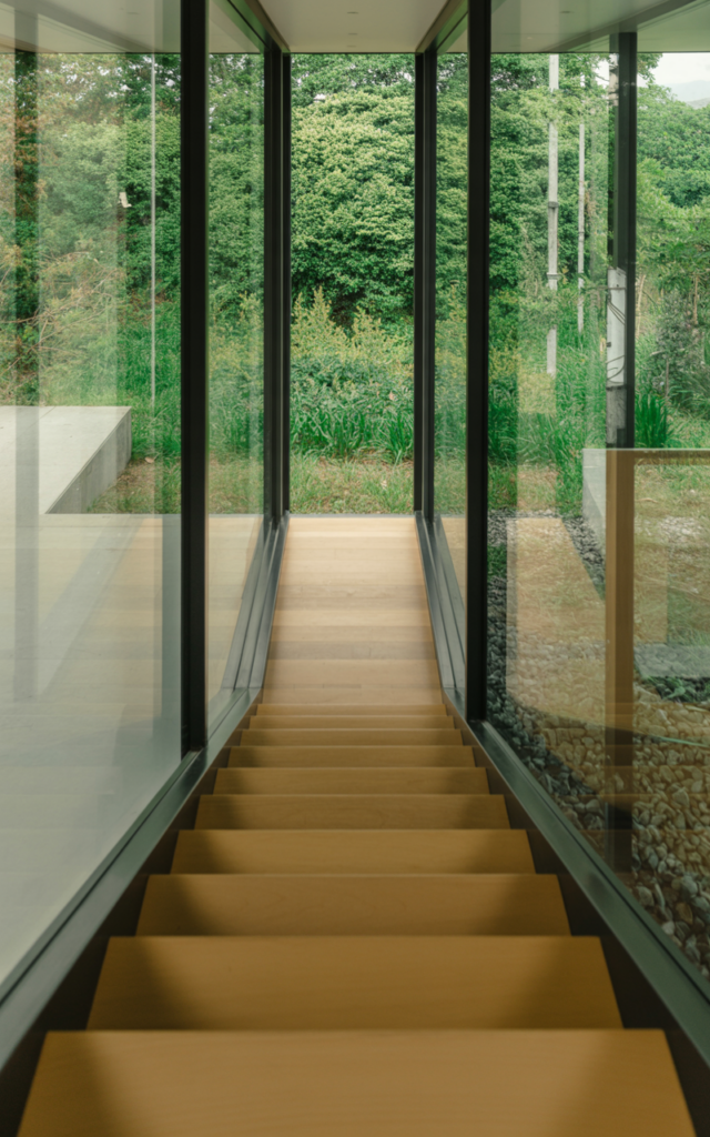 An elegant and natural-looking realistic photograph of a staircase with floor-to-ceiling glass panels running alongside the stair run, overlooking lush greenery with slim black frames and light wood treads decorated in modern minimalist style with natural lighting